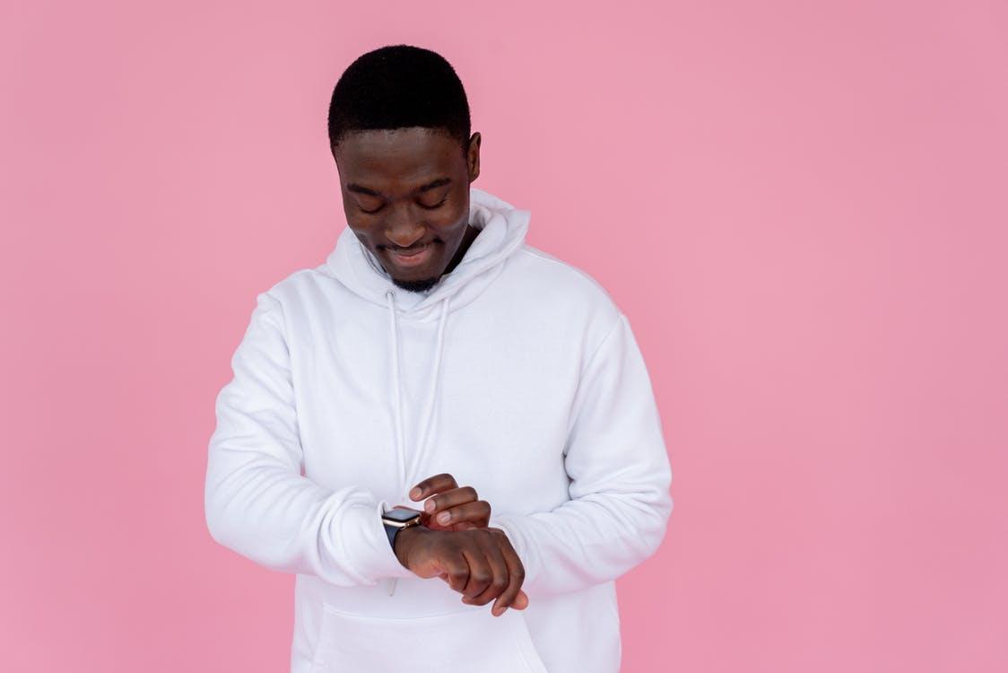 Smiling African American male in casual sweatshirt touching screen of modern smart watch while standing against pink background